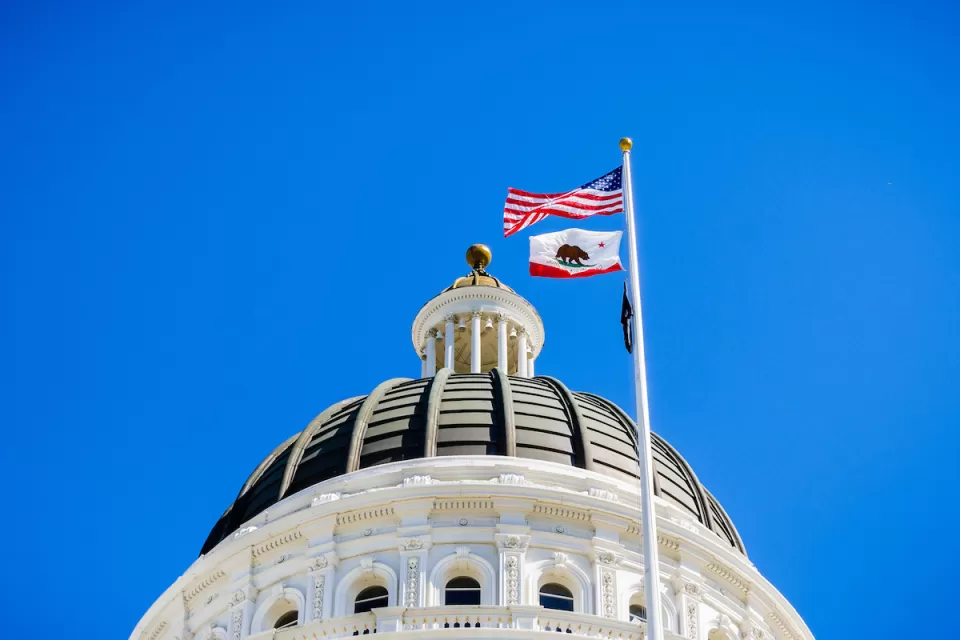 Close-up of top of California state capitol dome with U.S. and California flags flying and blue sky in background.